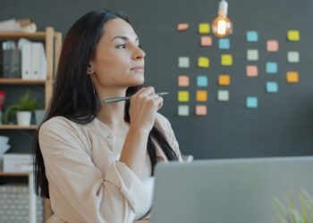 Beautiful young businesswoman is thinking then working with laptop in office, guy is busy in background. Ambitious people and occupation concept.