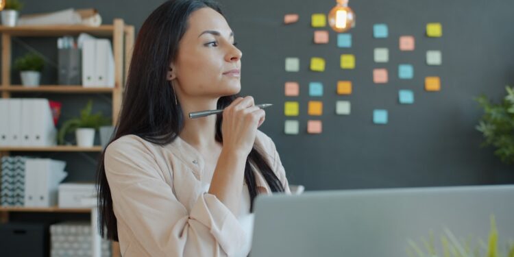 Beautiful young businesswoman is thinking then working with laptop in office, guy is busy in background. Ambitious people and occupation concept.