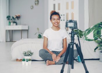 Good-looking African American girl creative blogger is recording video about plants sitting on floor of her modern apartment and talking looking at camera on tripod.