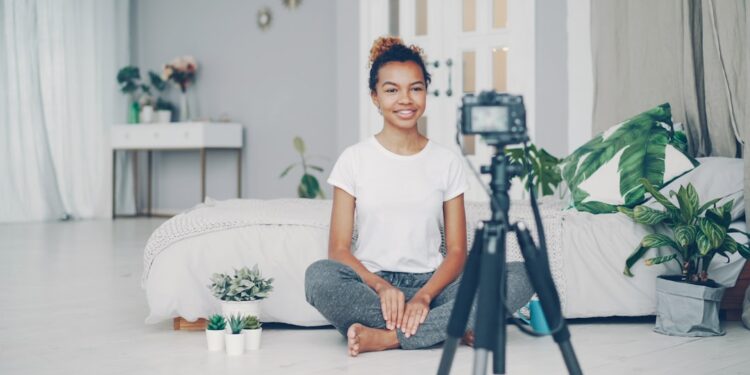 Good-looking African American girl creative blogger is recording video about plants sitting on floor of her modern apartment and talking looking at camera on tripod.
